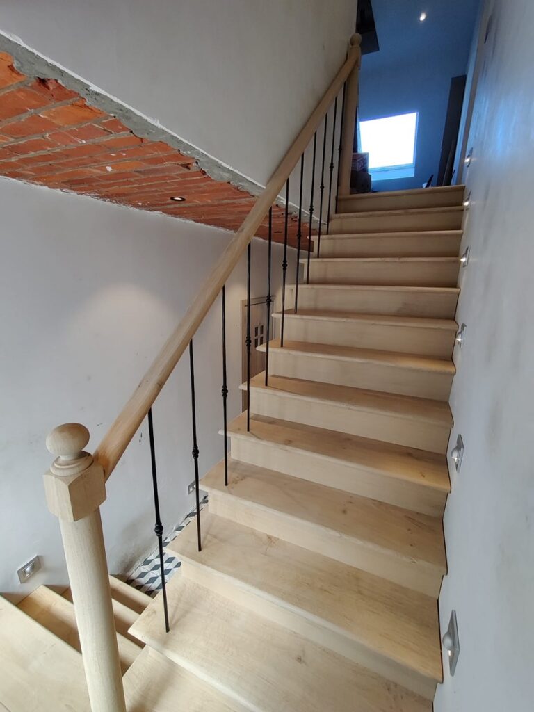 Landelijke trap in Engelse stijl tijdens plaatsing in Kalmthout, zicht op trappaal, handgreep en smeedijzeren balusters met zwarte poedercoating,treden met overstekende neus,treden die over de wangen komen,onbehandelde trap
